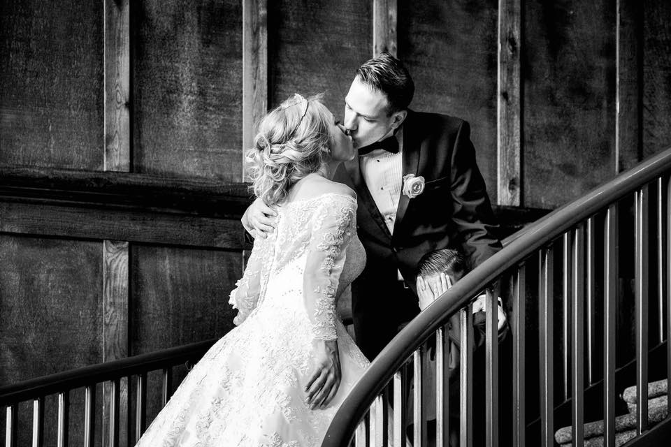 a black and white photograph of a bride and groom kissing on a grand staircase, with a little boy about the height of the railing holding his hands over his eyes