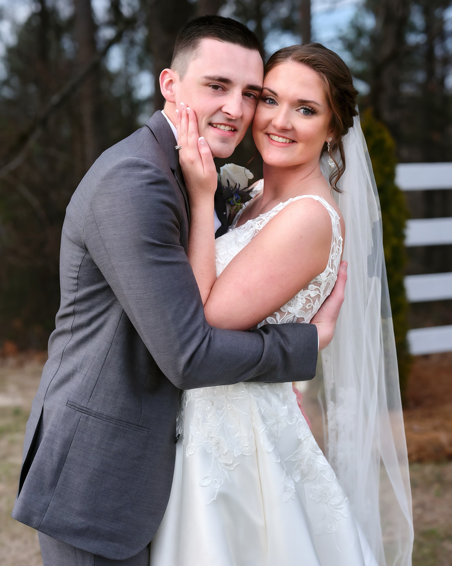 a young couple in an embrace, posing for a photograph, both smiling for the camera. The bride is holding the groom's face and the groom's arms are around her waist