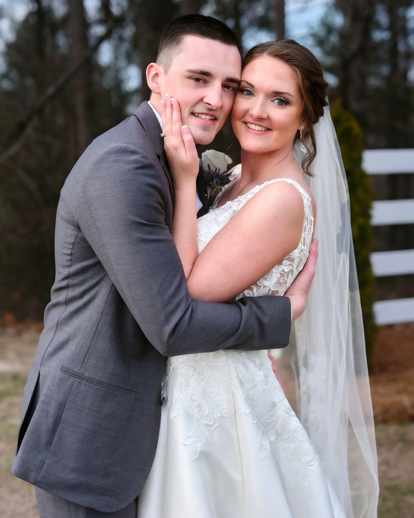 a young couple in an embrace, posing for a photograph, both smiling for the camera. The bride is holding the groom's face and the groom's arms are around her waist