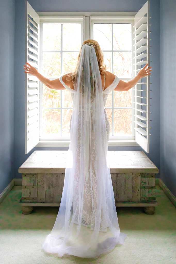 a photograph of a bride standing in a blue room, holding white open shutters, the bride is facing the window and we see the bride's dress and veil from the back only 