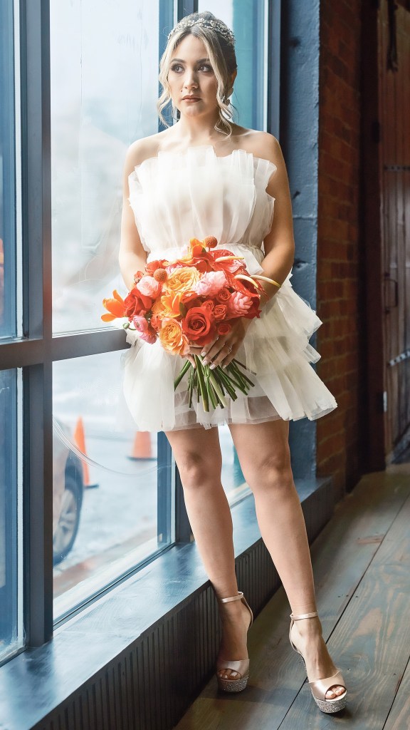 a bride standing by a grand window, looking out into the rain while holding a colorful red and orange bouquet of flowers