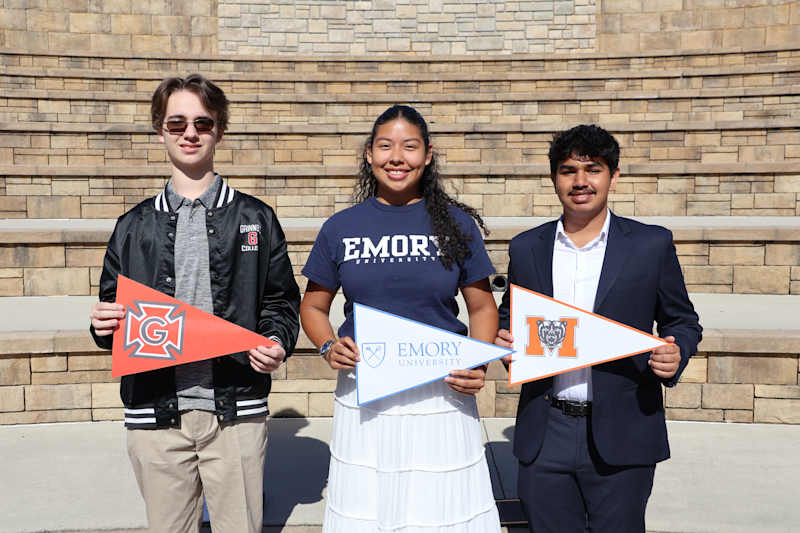 Three students stand, each holding a collegiate flag of the university he or she plans to attend.