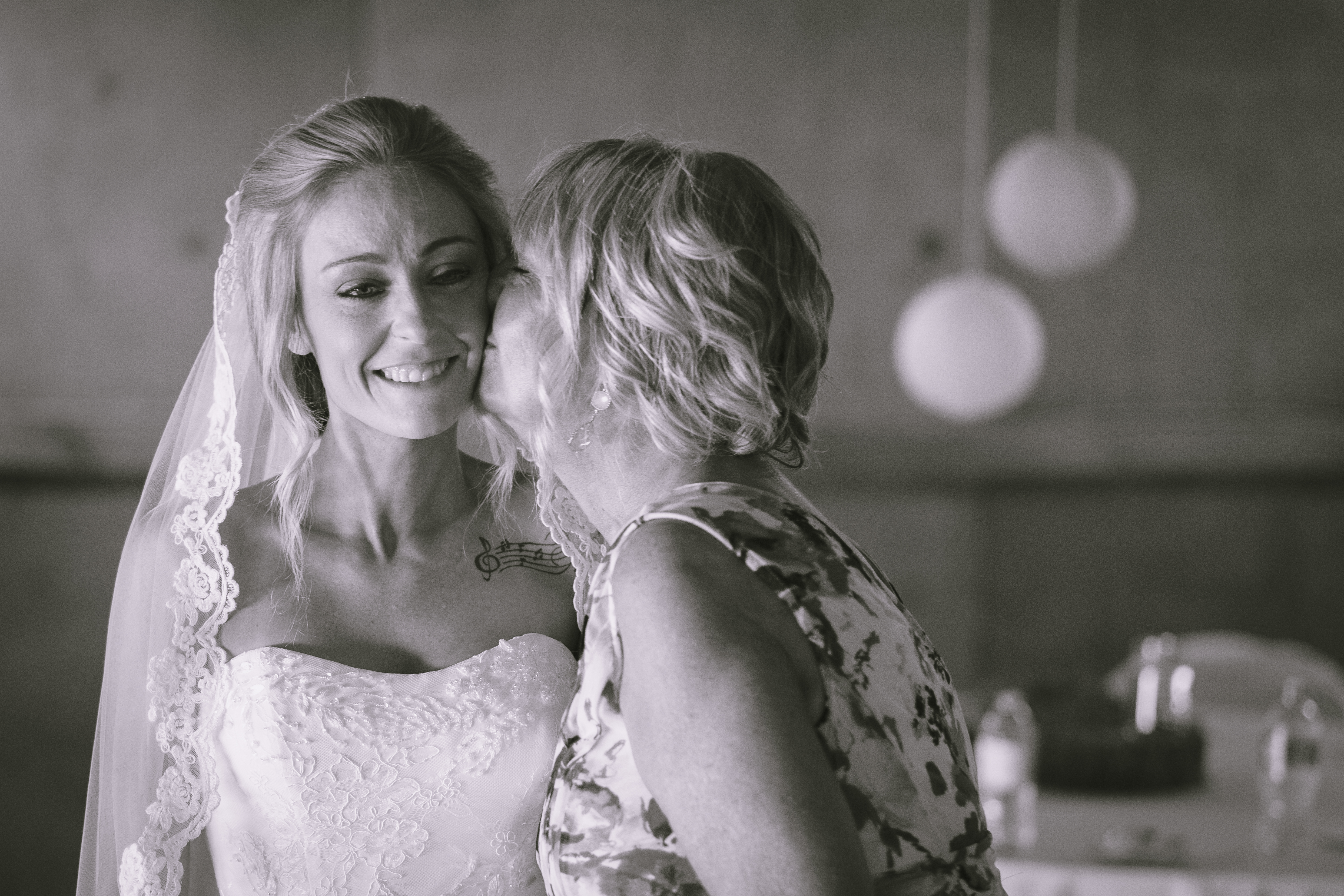 a bride dressed in her wedding dress, with mom kissing her on the cheek. photo is in black and white