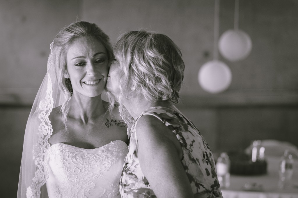 a bride dressed in her wedding dress, with mom kissing her on the cheek. photo is in black and white