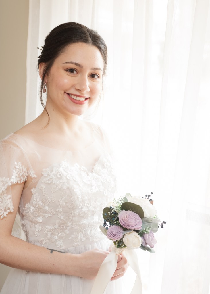 A bride stands by a cream colored walll and window holding a very small lavender bouquet